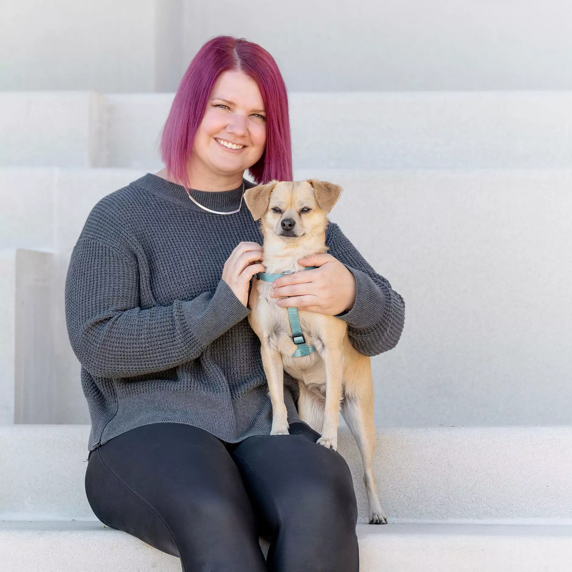 A white woman with magenta hair in a grey sweater sitting on steps. A small tan dog with floppy ears is confidently perched on her lap.